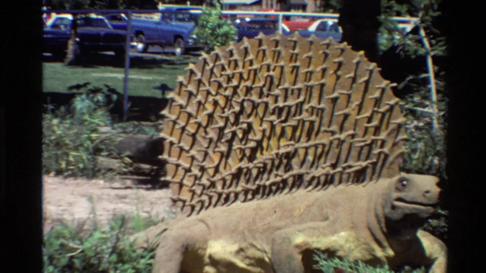 Home-movie view of the Stegosaurus statue from a similar angle, showing its plates, head, surrounding plants, fence, and background cars.