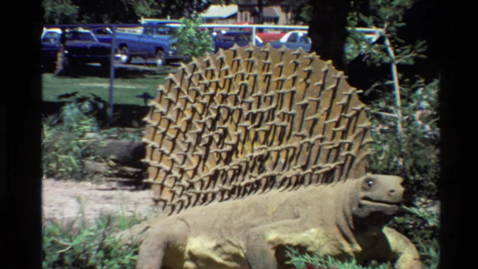 1980 home-movie frame centered on the Stegosaurus statue's textured plates and body, with fence, lawn, and parked cars visible behind.