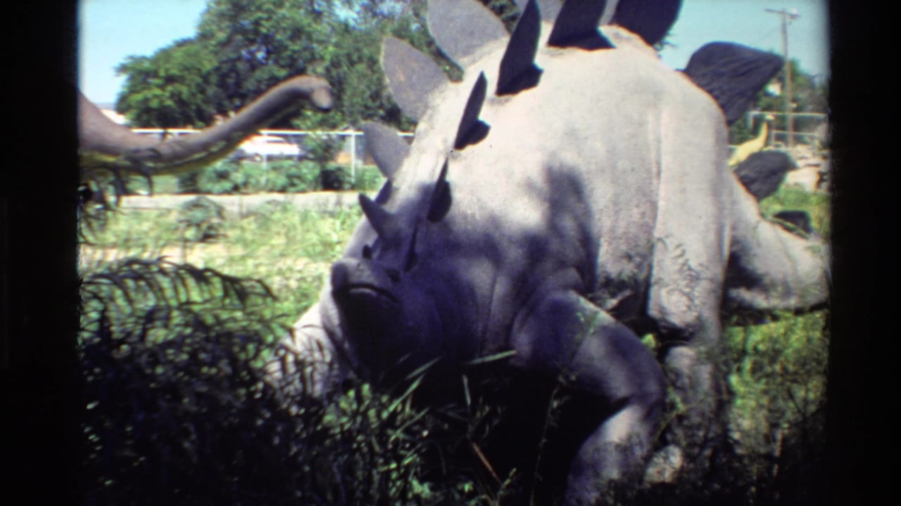 1980 Western USA 8mm frame showing a gray Stegosaurus model grazing among grass, with fencing, trees, and another dinosaur model in the background.