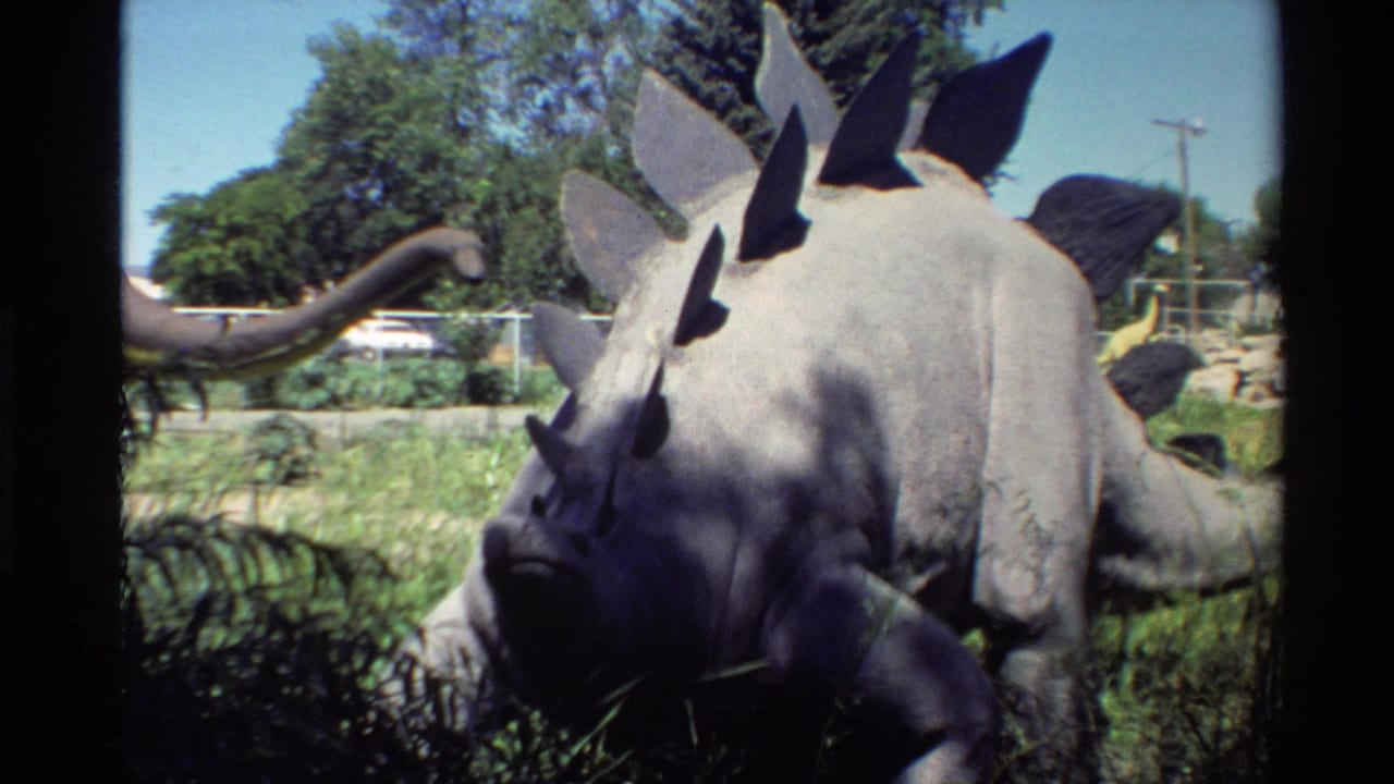 Stegosaurus model standing in grass with trees, fencing, and a long-necked model behind it.