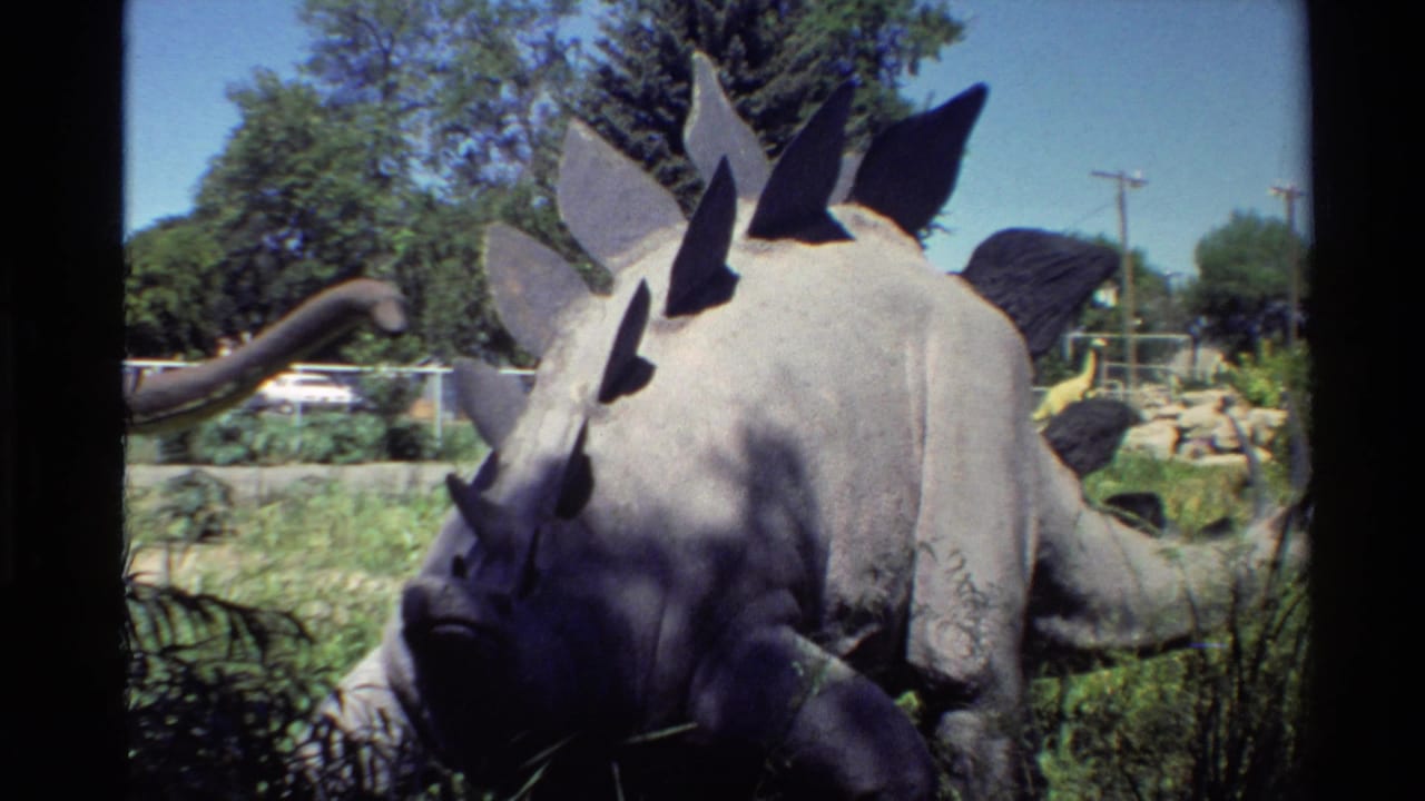 Outdoor 8mm frame of the Stegosaurus model from a slightly wider angle, showing grass, fencing, trees, and background exhibit figures.