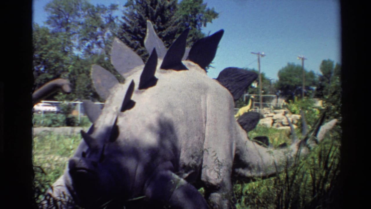 1980 Western USA 8mm frame showing a grey spiked dinosaur model in a landscaped outdoor park with trees and utility poles behind it.