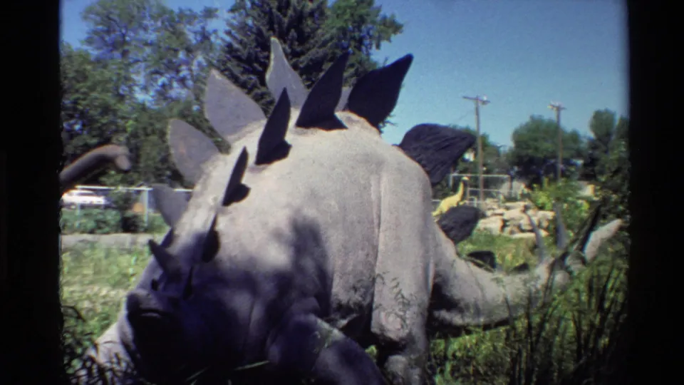1980 Western USA 8mm frame showing a grey spiked dinosaur model in a landscaped outdoor park with trees and utility poles behind it.