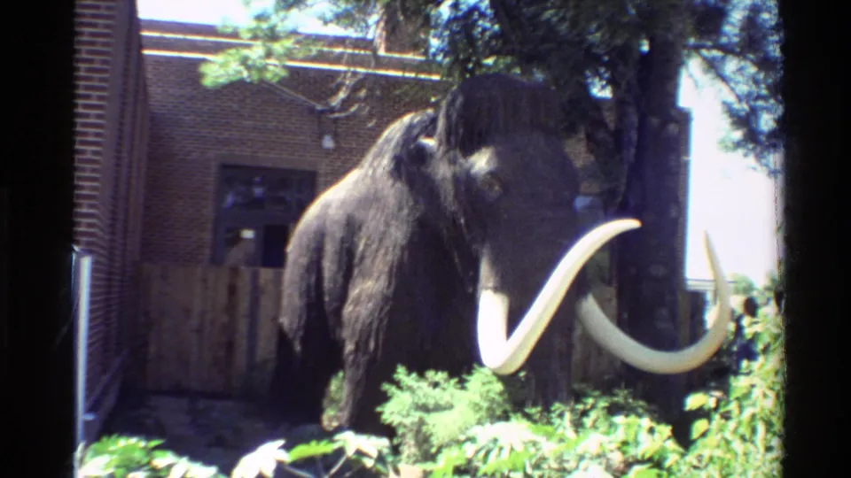 1980 Western USA home-movie frame of a mammoth statue in a zoo-like outdoor display with brick, fencing, and plants.