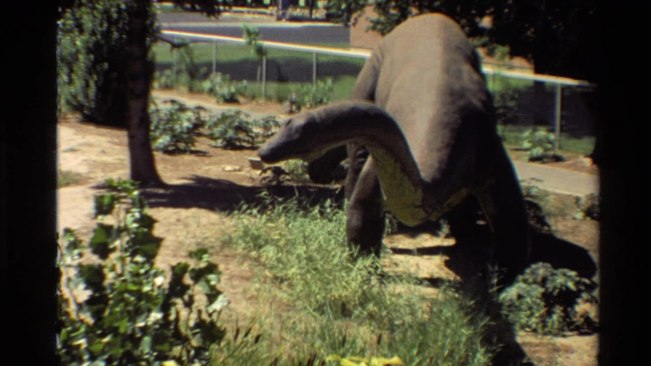 Large brontosaurus-style dinosaur model in a landscaped outdoor park area with grass, dirt, trees, and a fence visible.