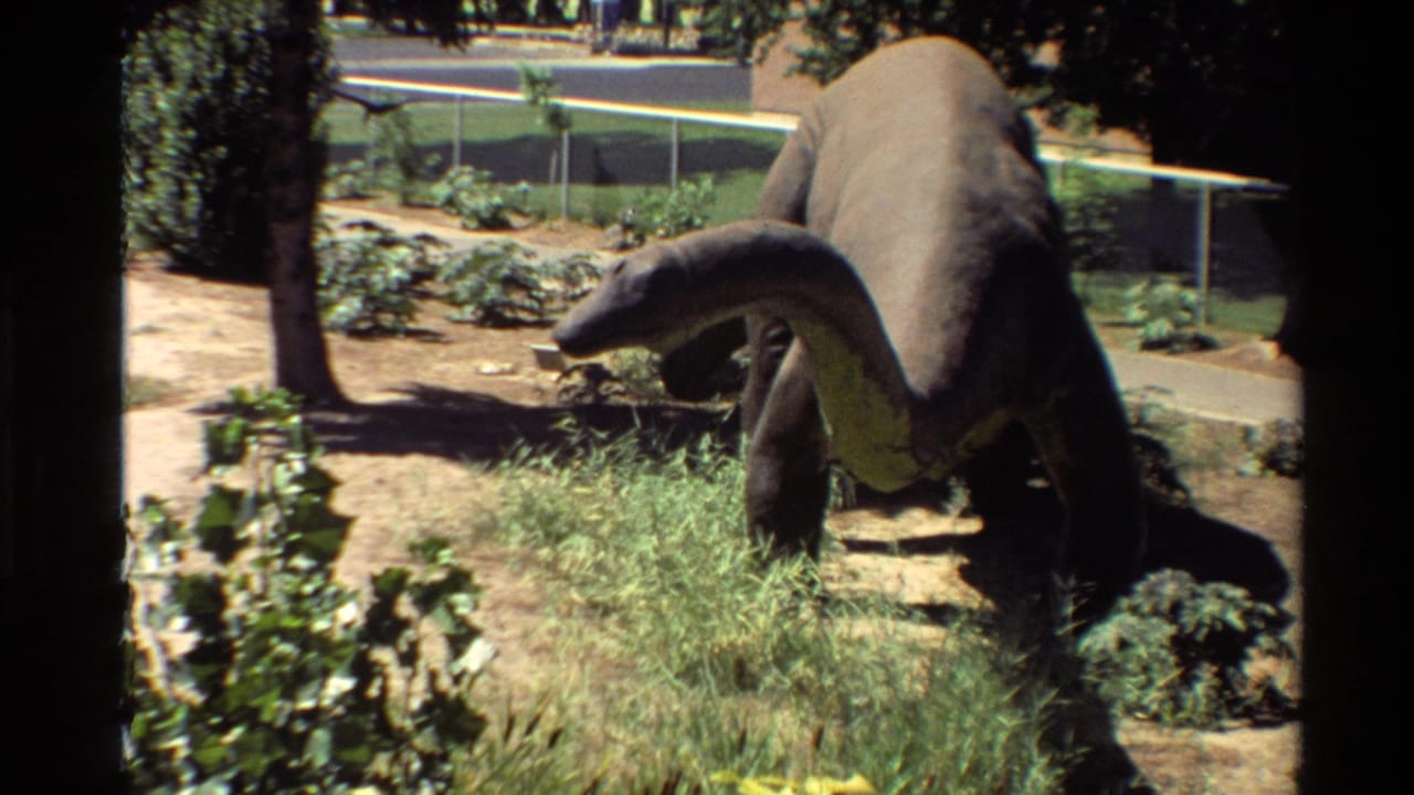 1980s 8mm frame of a large long-neck dinosaur statue in an outdoor U.S. park setting with grass, paths, trees, and chain-link fencing.