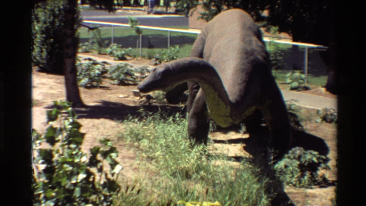 1980s 8mm view of a stationary long-neck dinosaur model in an outdoor park, with landscaped plants, fencing, and pavement behind it.