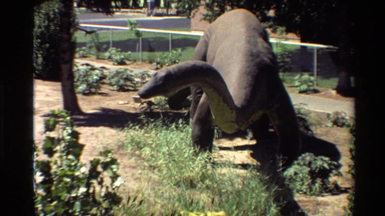 Vintage 8mm frame of a long-neck dinosaur statue in a sunlit outdoor exhibit area, with grass, dirt paths, trees, and chain-link fencing.