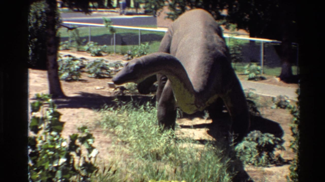 1980s U.S. home-movie frame showing a stationary long-neck dinosaur statue in a landscaped outdoor park with grass, trees, fencing, and distant structures.