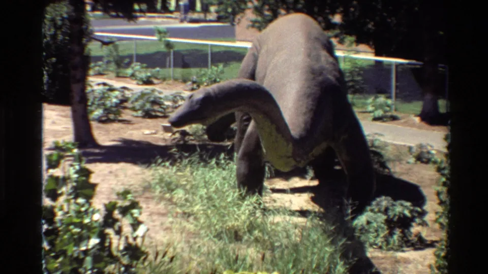 1980s U.S. home-movie frame showing a stationary long-neck dinosaur statue in a landscaped outdoor park with grass, trees, fencing, and distant structures.