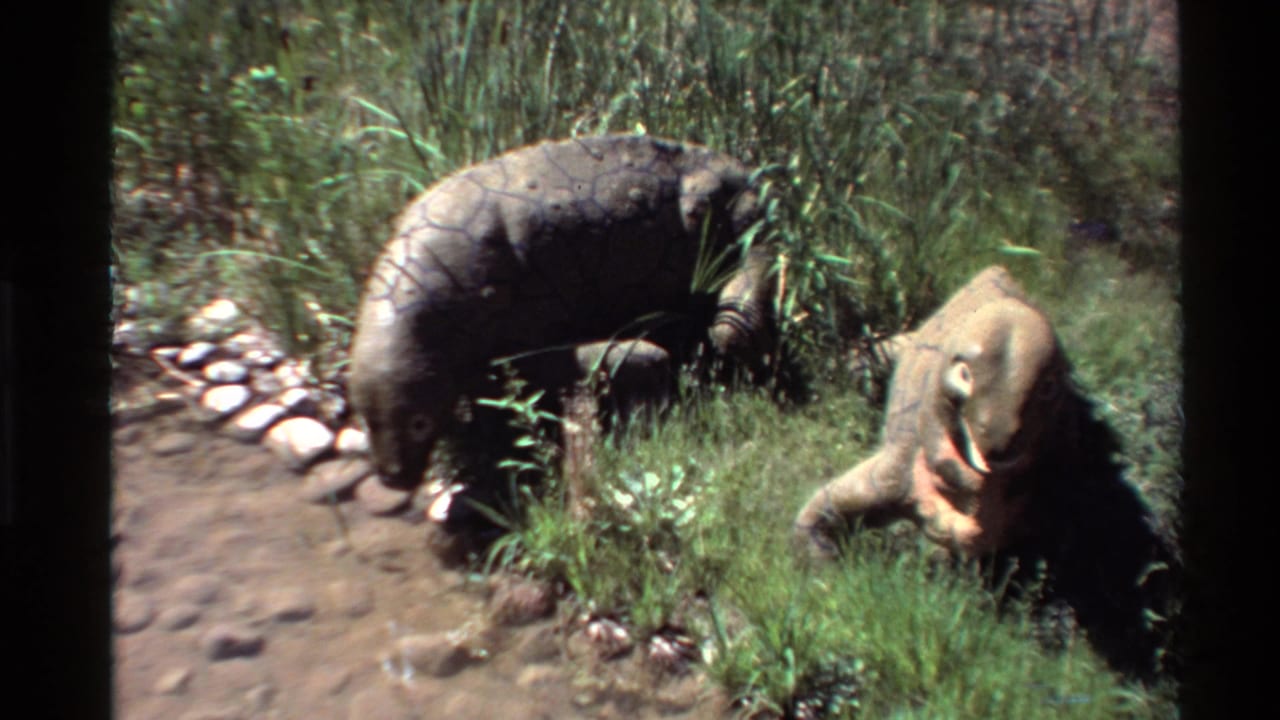 Large dark reptile-like animal bends near grass while a lighter animal rests nearby.