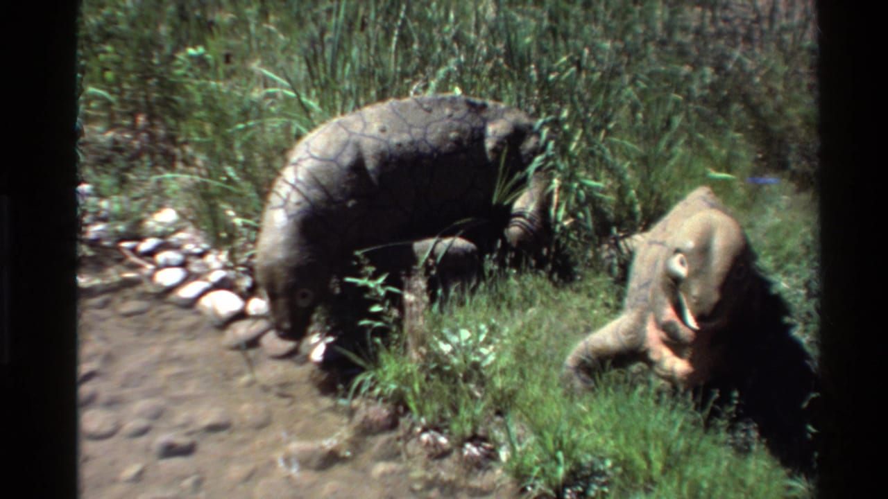 1980 Western USA 8mm frame showing grey and tan Ornithomimus models in a planted outdoor exhibit area.