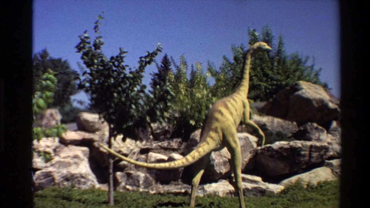 Yellow-brown dinosaur model standing among rocks and shrubs.
