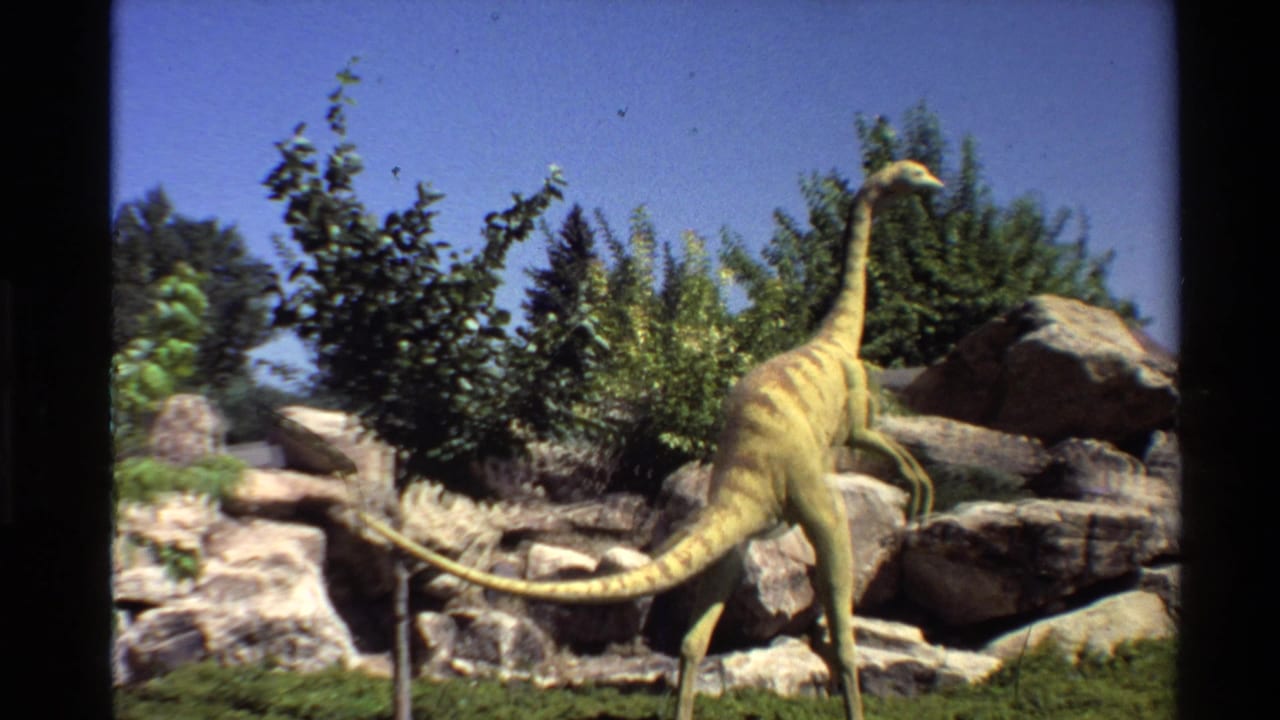 Final 8mm frame holds on the yellow-brown Ornithomimus model in the outdoor exhibit, with rocks, foliage, and clear sky visible.