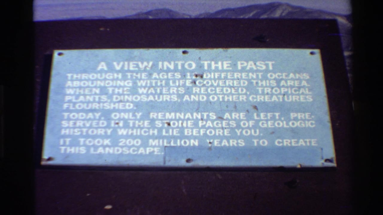 1980 Western USA home-movie frame showing a pale blue geologic-history plaque on a dark mount with distant mountains and sky behind it.