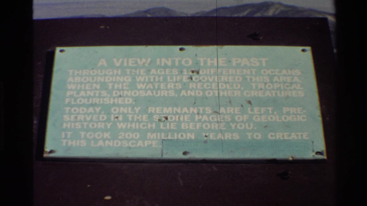 Visitor-shot 8mm frame of the geologic-history plaque, slightly tilted and softly exposed, with dark mount, screws and a narrow mountain backdrop.