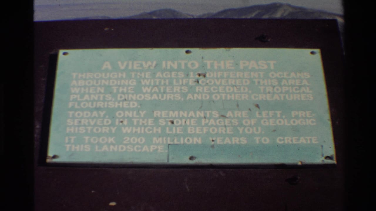 Final 8mm frame of the Western USA geologic-history plaque, showing faded white text, screw fasteners, dark mounting surface and distant mountains.