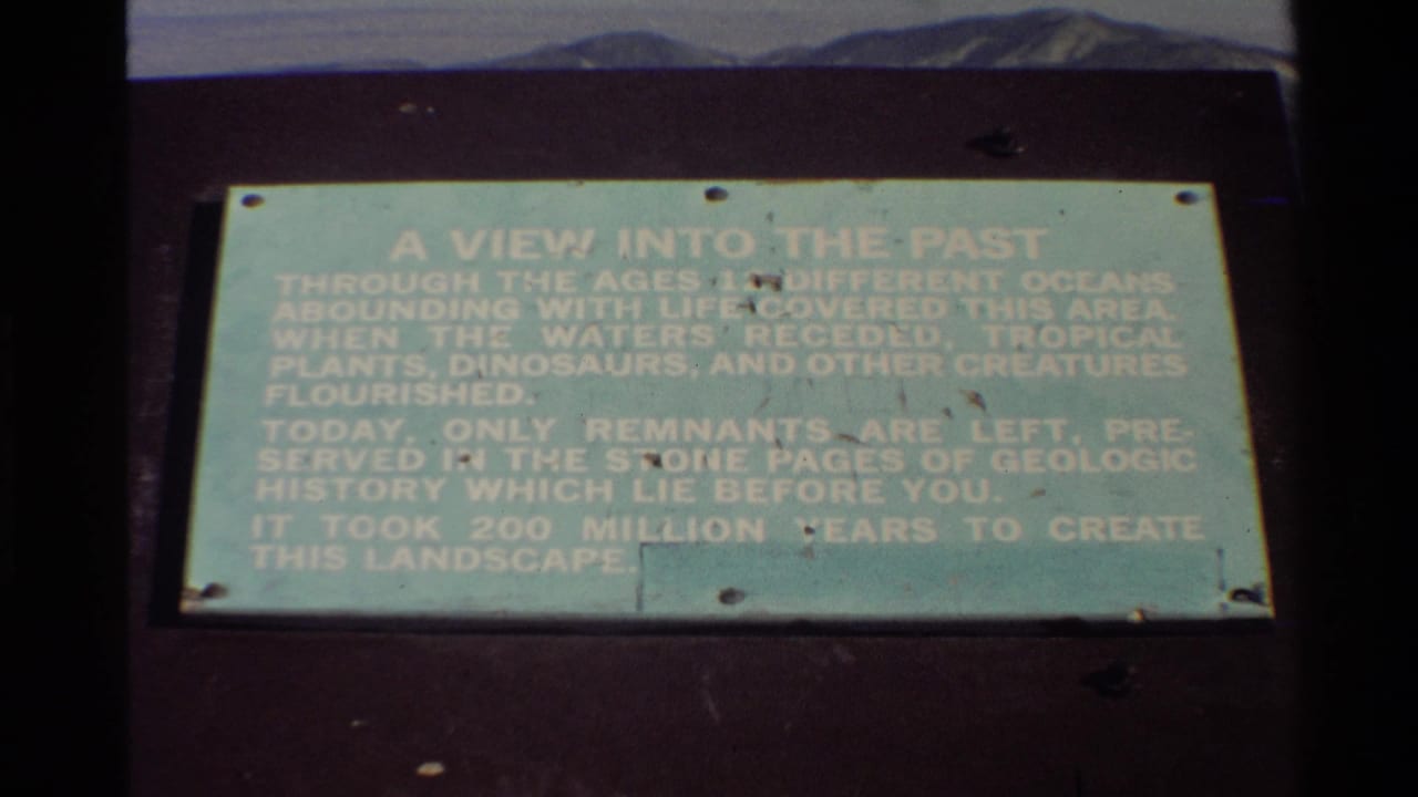 1980 Western USA 8mm frame showing a pale green geologic history sign on a dark mount, with distant hills and sky above.