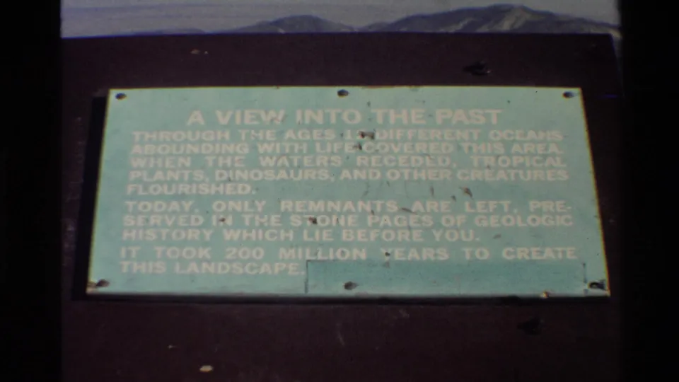 1980 Western USA 8mm frame showing a pale green geologic history sign on a dark mount, with distant hills and sky above.
