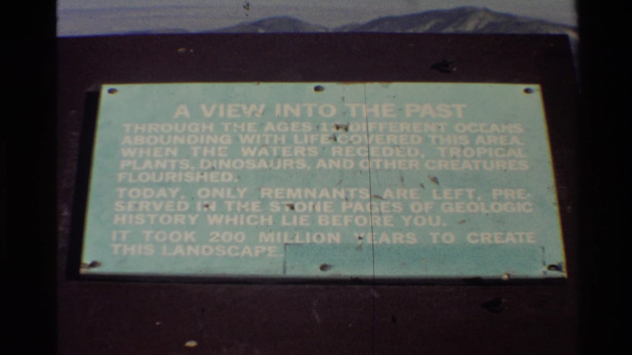 Pale green outdoor display sign with white text, dark support, distant hills, and film scratches.