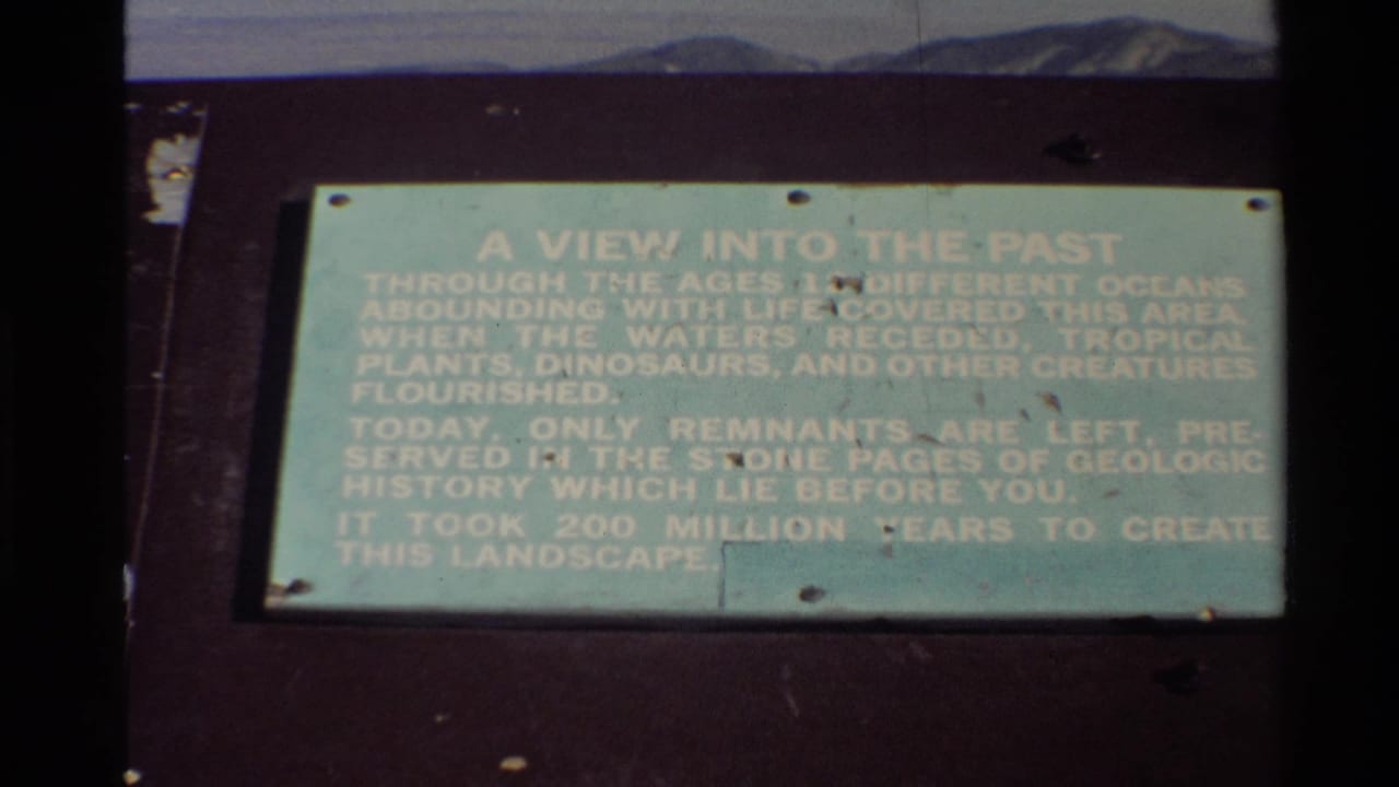 Visitor-shot 1980 8mm frame of a weathered geologic-history plaque with text about ancient oceans, plants, and dinosaurs.