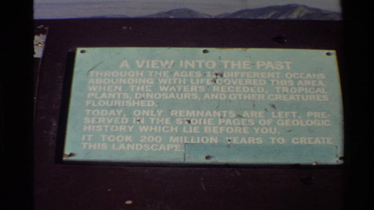 1980s 8mm frame of a geological interpretive sign titled A View Into The Past, filmed close up with dark film borders and distant terrain behind it.