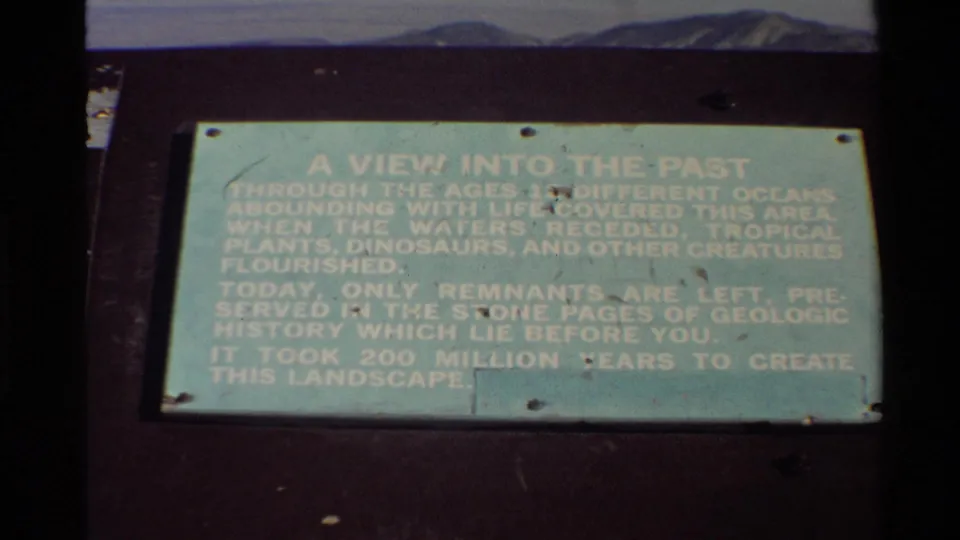 1980s 8mm frame of a geological interpretive sign titled A View Into The Past, filmed close up with dark film borders and distant terrain behind it.