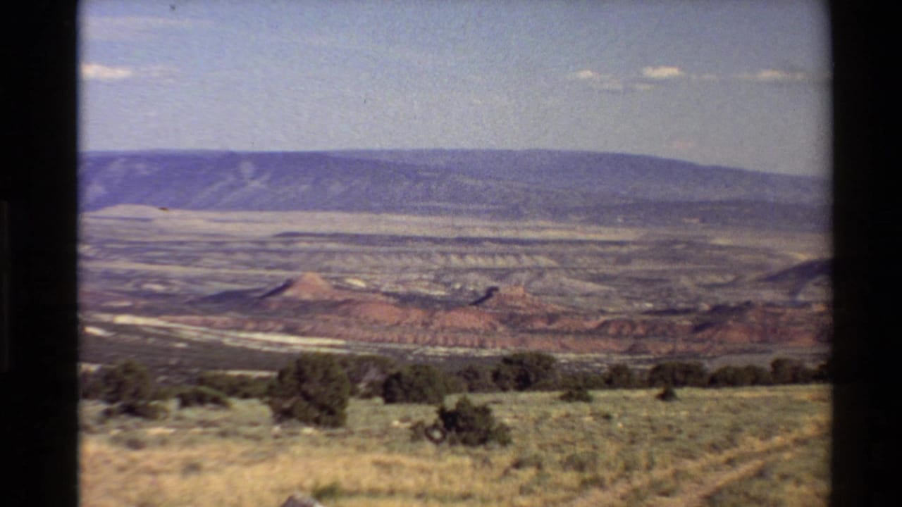 Wide 8mm view of an arid Western U.S. landscape with scrubland, layered red rock, broad mesas, distant mountains, and pale sky.