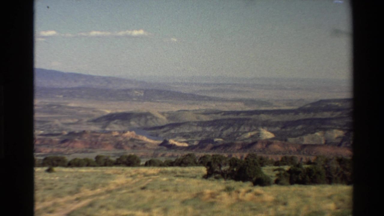 1980 home-movie frame from a high overlook, with dry grass in front of red-toned hills and hazy Western U.S. landforms beyond.