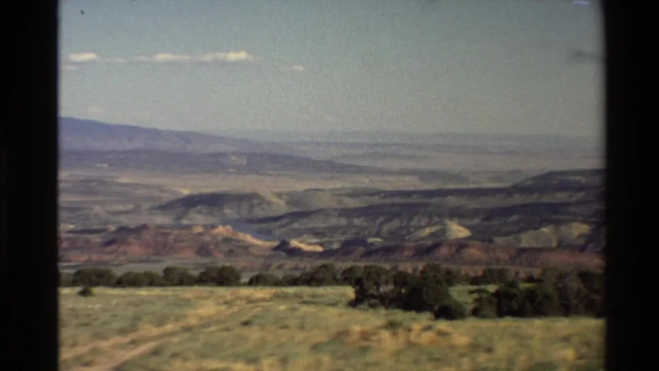 1980 home-movie frame from a high overlook, with dry grass in front of red-toned hills and hazy Western U.S. landforms beyond.