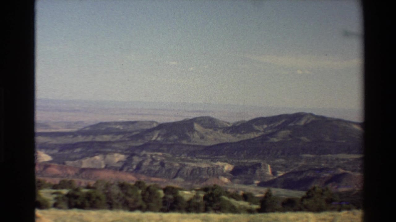 Western USA 1980 home-movie frame with dark trees, dry meadow grass, and layered hills receding into haze.