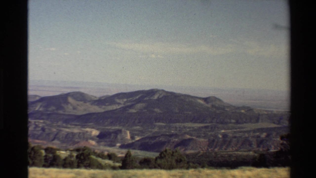 1980 8mm Western U.S. home-movie frame showing grassy land, scattered trees, ridges, haze, and a pale blue sky.