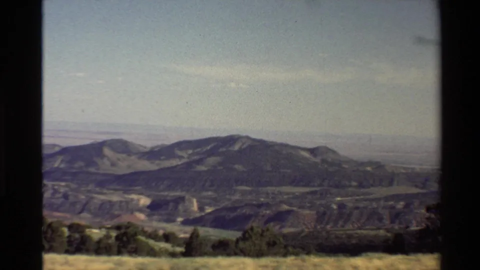 1980 8mm Western U.S. home-movie frame showing grassy land, scattered trees, ridges, haze, and a pale blue sky.