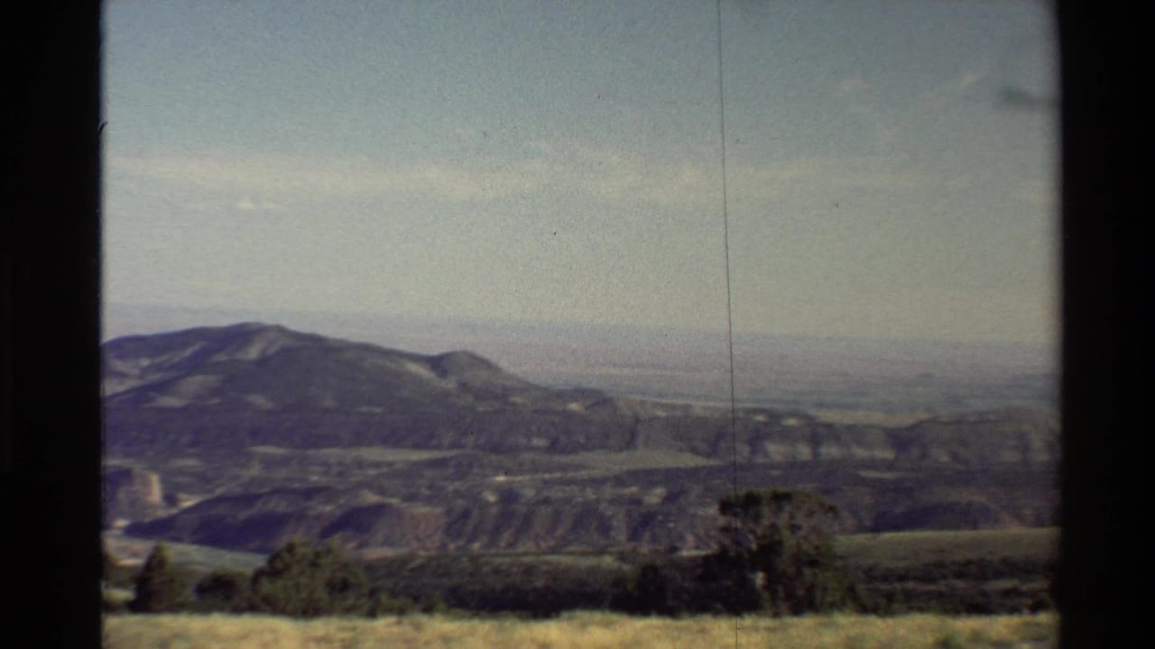 Soft 1980 8mm frame of Western U.S. mountains, open valley, dry grass, shrubs, and pale sky from a travel viewpoint.