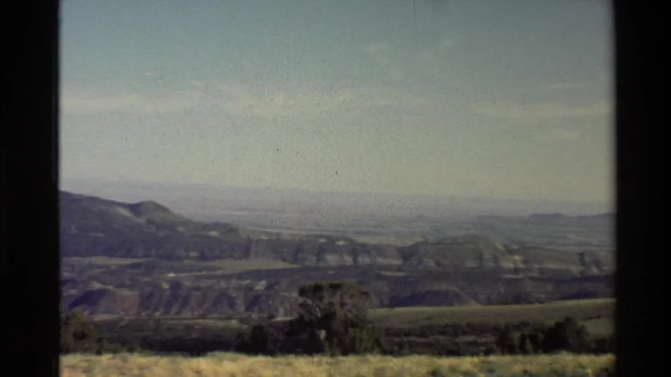 Western U.S. 1980 home-movie still showing low vegetation, rolling mountain ridge, distant plain, haze, and light blue sky.