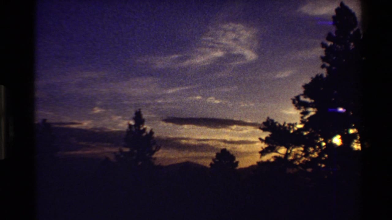 Supplied 1980 Western USA 8mm frame showing silhouetted pines and distant hills beneath a grainy sunset sky with clouds and warm light at right.