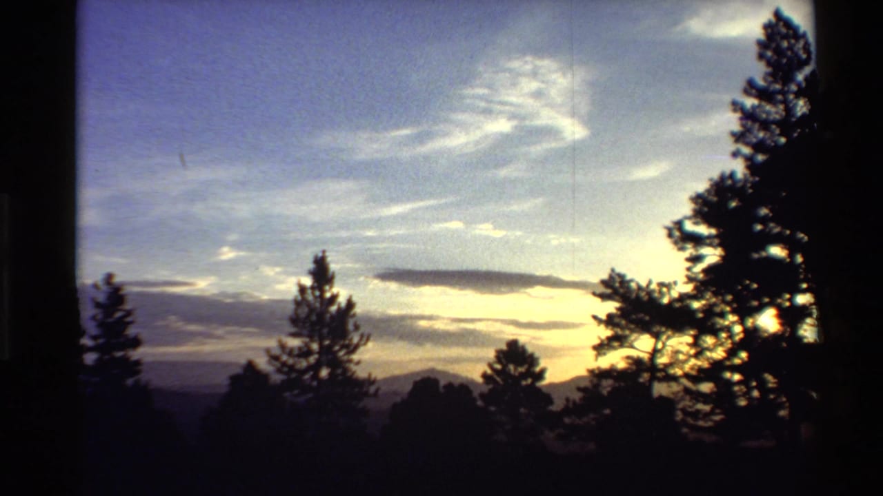 Brighter 8mm frame from the supplied 1980 Western USA clip with pine silhouettes, distant hills, pale clouds, and low sunlight glowing at right.