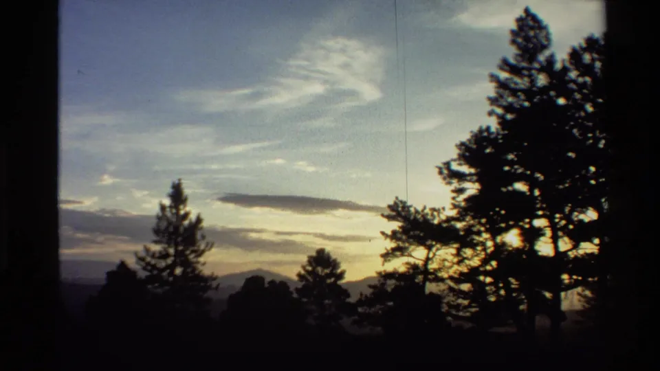 1980 Western U.S. home-movie frame with pine trees in silhouette, wispy clouds, and dim sunset light near the horizon.