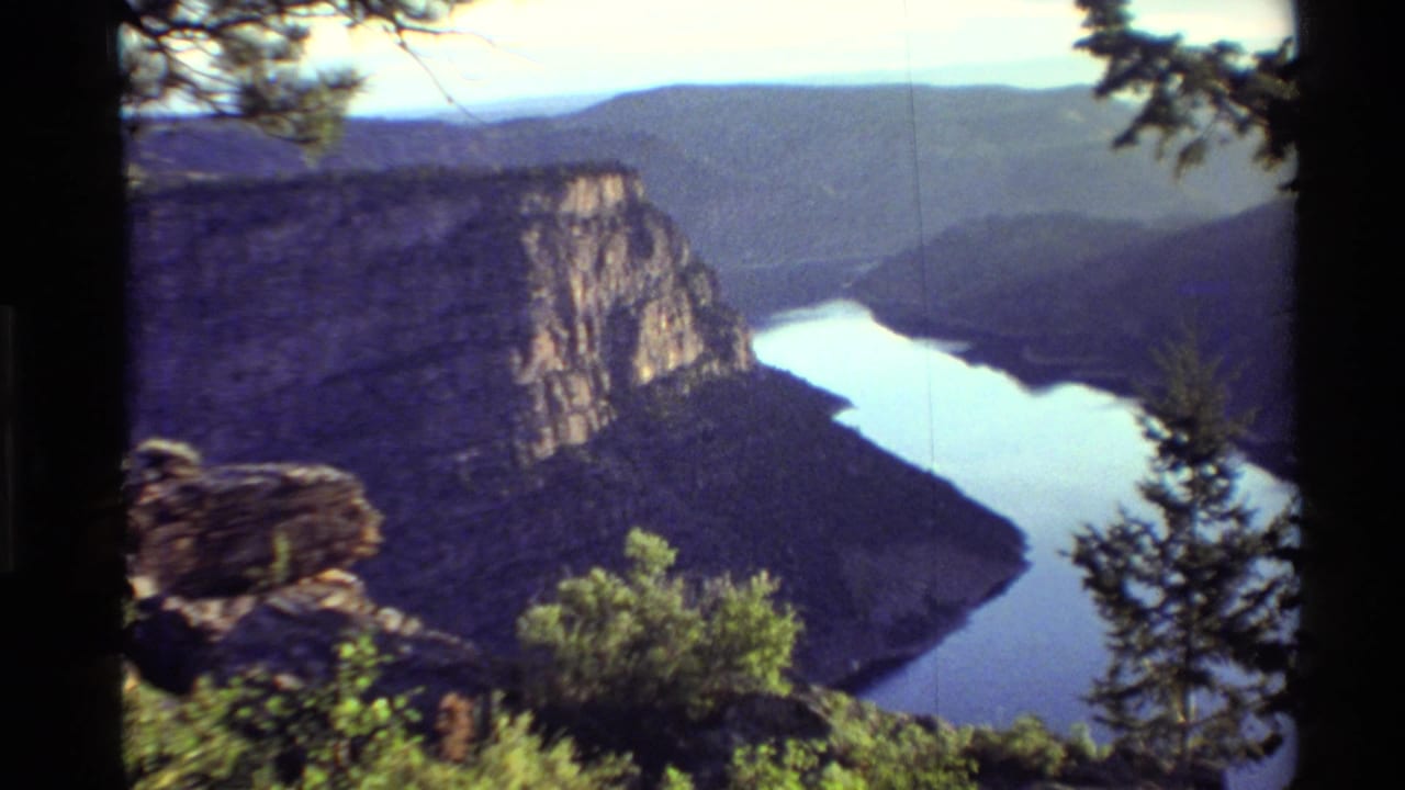 High 8mm home-movie view of a Western USA river canyon, with a cliff wall, shadowed hills, and foreground brush.