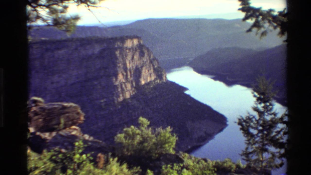 1980 Western USA 8mm view from a high canyon overlook, showing a winding river, sheer rock wall, foreground trees, and hazy hills.