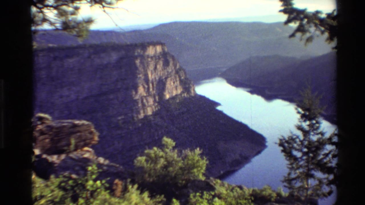 Canyon and river framed by dark tree branches, with a rocky cliff and layered hills beyond.
