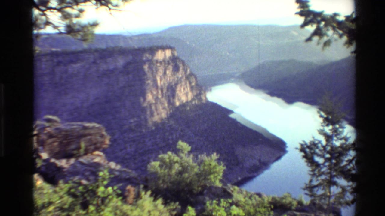 Western USA 1980 home-movie frame showing a bright river through a deep canyon, with cliff face, trees, shrubs, and hazy hills.