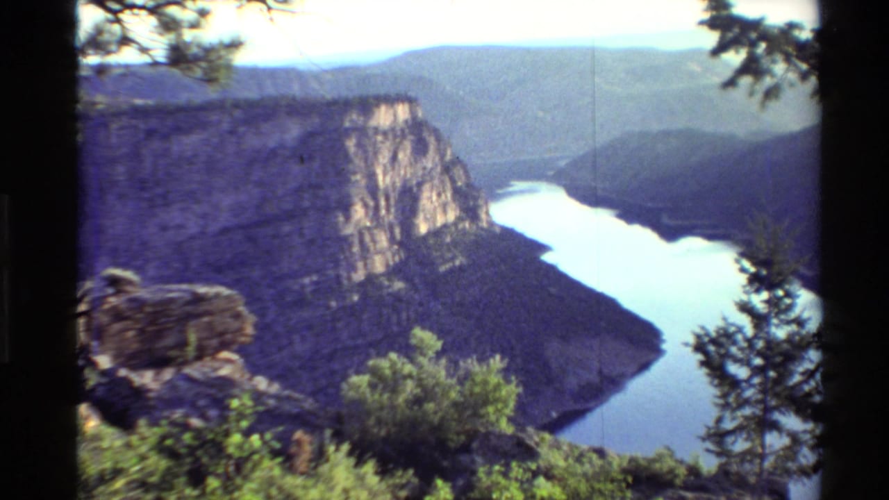 1980s 8mm Western U.S. overlook view with a winding river below steep canyon walls, framed by pine branches and foreground shrubs.