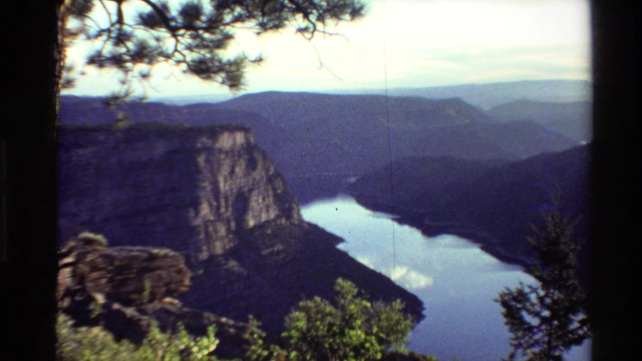 Soft 1980s 8mm landscape frame with a canyon river below, rocky overlook ledge, pine tree, green shrubs, and layered Western U.S. hills.