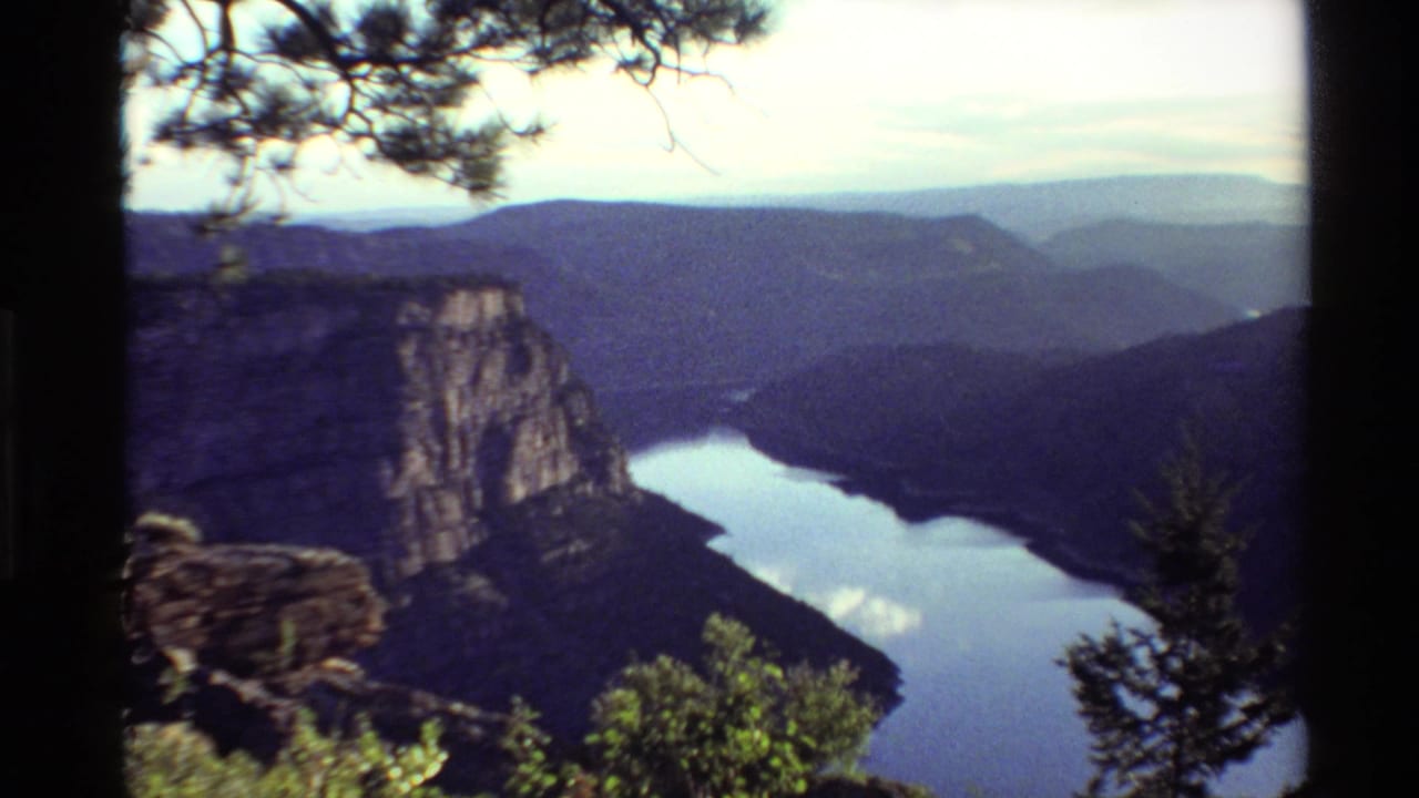 1980s 8mm Western U.S. canyon vista seen from a shaded overlook, with river water, distant ridges, pine branches, and shrubs.