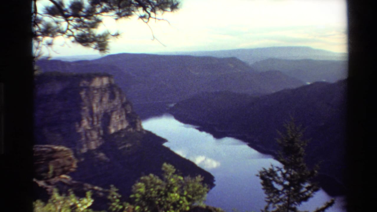 Soft 8mm landscape frame of a canyon lake, rocky wall, pine branches, and distant blue ridgelines in the Western United States.