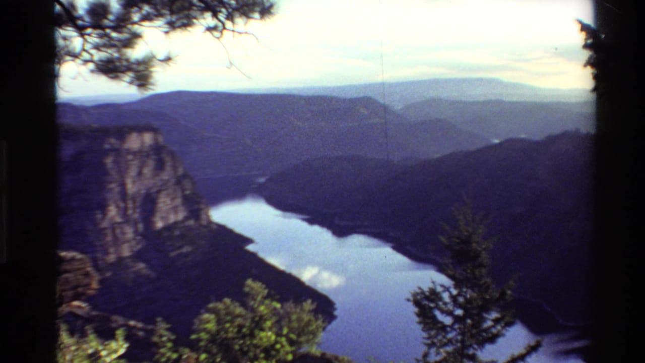 Vintage 8mm overlook view of a lake between canyon walls, with shrubs in front and hazy mountain ridges beyond.