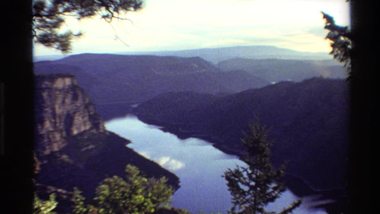 Faded-color 8mm frame of a Western United States canyon lake vista with cliffs, pine silhouettes, and distant hills.