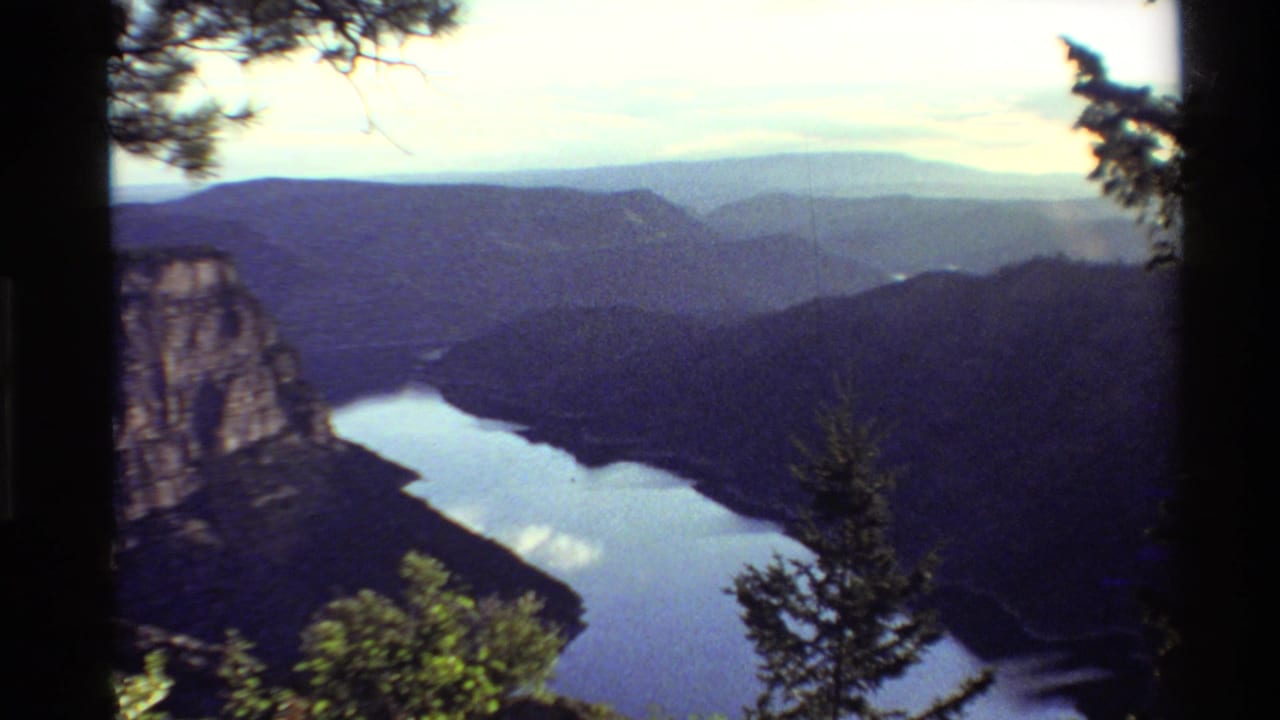 1980 Western USA 8mm view of a mountain valley and reflective lake framed by trees, with layered ridges under a pale sky.