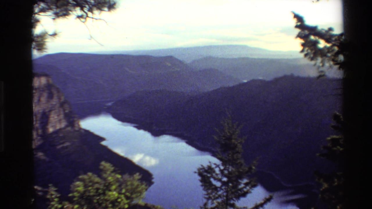 1980 Western USA home-movie frame showing a reflective lake in a mountain valley, bordered by forested ridges and foreground branches.